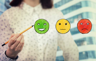 Image of a woman holding a pencil to a green smiley face. There is also a yellow face and mad red face
