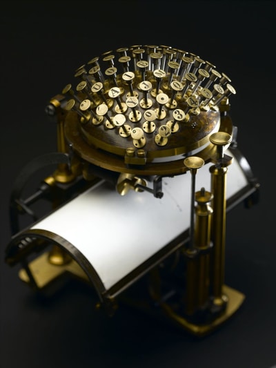 The Hansen Writing Ball has brass colored keys arranged as if on the top half of a ball, with a curved sheet of paper resting under them.