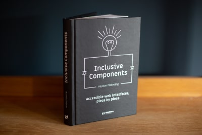 A picture of a black-and-white book cover standing on a wooden surface abd tilted to the side titled Inclusive Components, Accessible web interfaces, piece by piece, written by Heydon Pickering
