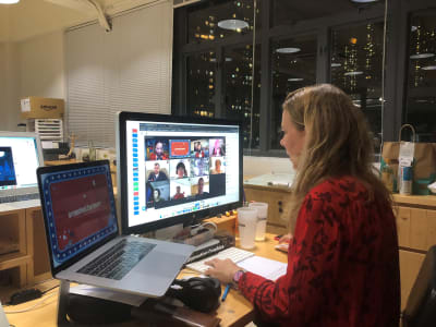 A woman sitting at two screens, one has lots of people on video